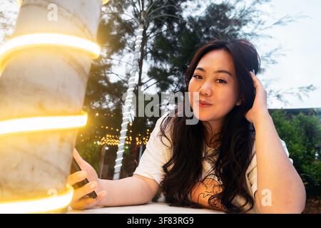 junge asiatische Frau, die die Kamera mit einem subtilen Lächeln ansieht, während sie abends im Freien sitzt und ein Smartphone in der Nähe von warmer dekorativer Beleuchtung hält Stockfoto