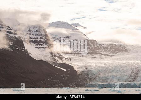 Nebel zieht über Schneefelder und Felsen, während ein Gletscher im arktischen Licht von Borgvig in Grönland auf das Wasser zufließt Stockfoto