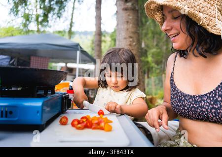 junge Frau mit Kleinkind-Tochter kocht zusammen als Familie auf dem Campingplatz mit Gaskampferherd im Freien, Sommerurlaub-Lebensstil Stockfoto