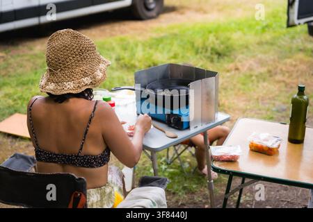 junge Millennials-Frau, die auf einem Campingplatz mit einem Gas-Campingkocher im Freien in der Natur kocht, mit Hut im Sommer und Zelt im Hintergrund Stockfoto