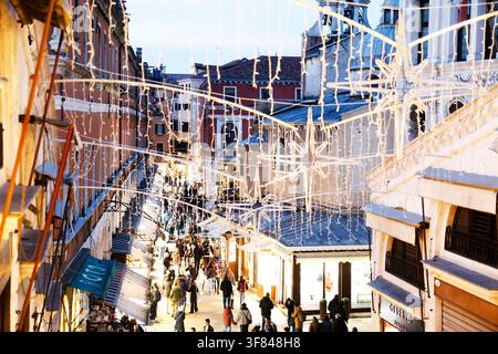 Straßenszene in der Dämmerung in Venedig mit Weihnachtslichtern und lebhaften Menschenmassen auf engen Wegen, die mit Geschäften gesäumt sind Stockfoto