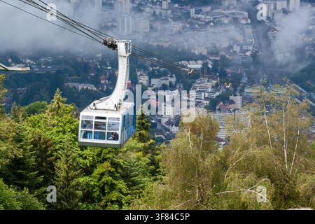 Seilbahngondel bewegt sich über einen üppigen grünen Wald mit Blick auf die Stadt unter bewölktem Himmel, Österreich Stockfoto