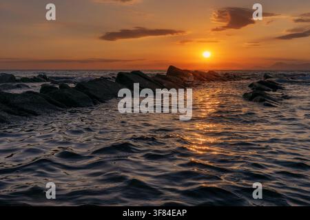 Scenischer goldener Sonnenuntergang über einer felsigen Seeküste mit Sonnenreflexion auf kräuselnden Wasserwellen. Stockfoto