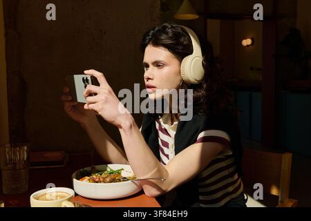 Lässiges Mittagessen, sie genießt das Essen, während sie in ein modernes Café vertieft in ihr Handy schaut Stockfoto