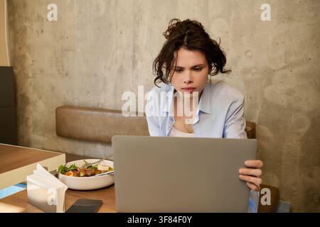 Frau mit lockigem Haar vertieft in ihren Laptop, während sie ihr Mittagessen in einem gemütlichen Café genießt Stockfoto