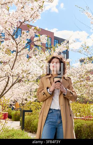 Stylische junge Frau in einem Trenchcoat genießt Kaffee, umgeben von blühenden Bäumen Stockfoto
