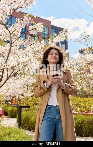 Eine modische junge Frau genießt Kaffee im Freien, umgeben von blühenden Kirschbäumen Stockfoto