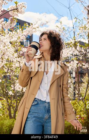 Charmante junge Frau in einem beigen Trenchcoat genießt Kaffee neben blühenden Kirschbäumen Stockfoto