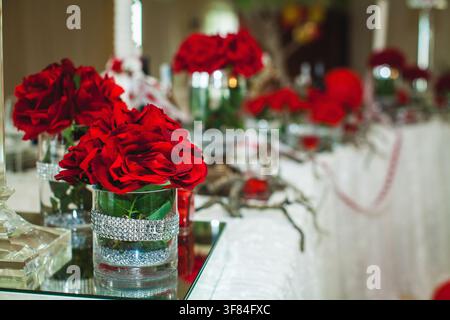 Ein Hochzeitstisch ist elegant mit roten Rosen in Glasvasen, Kerzen und einem zentralen Blumenarrangement dekoriert. Stockfoto