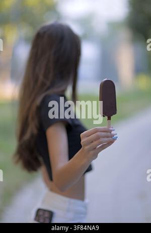 Junge Frau hält eine erfrischende Schokoladen-Eisriegel am Stiel und genießt einen süßen Snack an einem warmen Sommertag in einer Parkumgebung Stockfoto