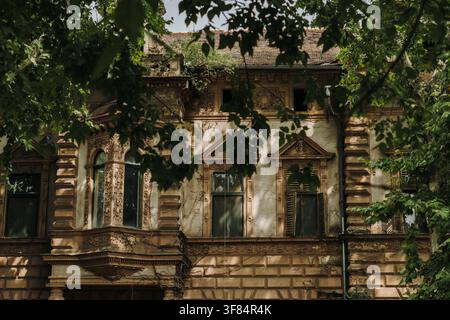 Verwitterte historische Villa teilweise von üppigen Bäumen und Laub verdeckt. Verlassene Architektur mit rissiger Fassade, die eine mysteriöse Atmosphäre schafft. Stockfoto