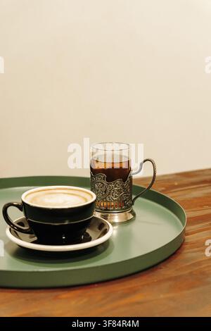 Frischer Cappuccino und schwarzer Tee, die zusammen auf einem modernen grünen Café-Tisch serviert werden. Gemütliche Café-Atmosphäre mit minimalistischer Komposition und Kopierplatz. Stockfoto