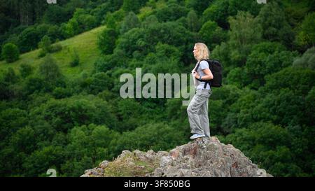 Eine kaukasische Frau mit blonden Haaren steht im Profil auf einem Felsvorsprung und blickt über eine üppige, grüne Landschaft. Sie trägt ein hellgraues T-Shirt Stockfoto