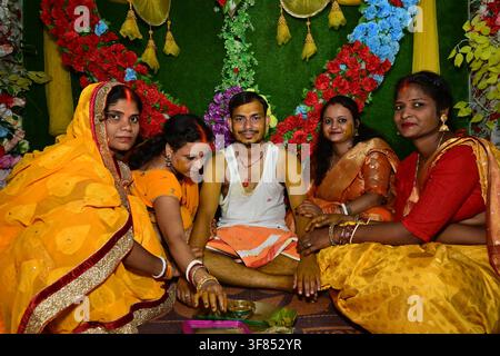 Indischer Mann in weißer Weste und orangefarbenem Dhoti sitzt mit Frauen in gelb-orangen Saris, die Haldi auftragen, während einer traditionellen Haldi-Zeremonie mit floralem Herzba. Stockfoto