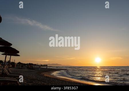 Goldener Sonnenuntergang beleuchtet Meereswellen an einem sandigen Strand. Malerische Küstenlandschaft mit Strohdächern, Liegestühlen, dunklen Wasserreflexionen und fernen Hügeln Stockfoto