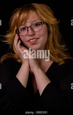 Junge Frau mit erdbeerblonden Haaren und Brille lächelt vor schwarzem Hintergrund Stockfoto
