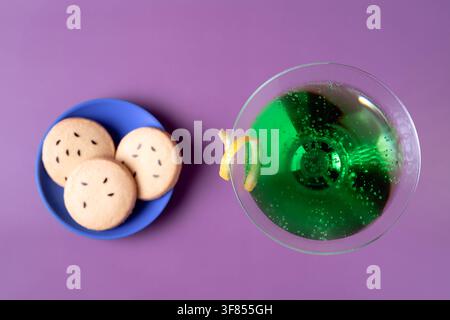 Überkopf-Flachlage eines grünen Halloween-Martini-Cocktails, garniert mit Zitronenschale, neben Fledermauskeksen vor einem violetten Hintergrund mit sauberem Kopierraum Stockfoto