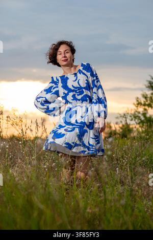 Vollständige Portrait einer Frau in einem blau gemusterten Sommerkleid, die bei Sonnenuntergang auf einer grasbewachsenen Wiese steht und Selbstvertrauen und unbeschwerte Freude ausstrahlt. Stockfoto