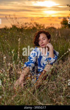 Neugierige Frau in blau gemustertem Kleid sitzt im hohen Gras zur goldenen Stunde, entspanntes Porträt in einer natürlichen Wiese. Stockfoto