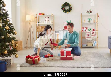 Fröhliches Paar sitzt auf einem Teppich in ihrem gemütlichen Wohnzimmer und tauscht an Weihnachten Geschenke aus. Stockfoto