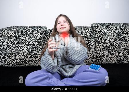 Krankes Mädchen sitzt im Schneidersitz auf dem Sofa, hält ein Glas Wasser und berührt ihren wunden Hals mit roter Hervorhebung für Halsschmerzen und Erkältungskonzept Stockfoto
