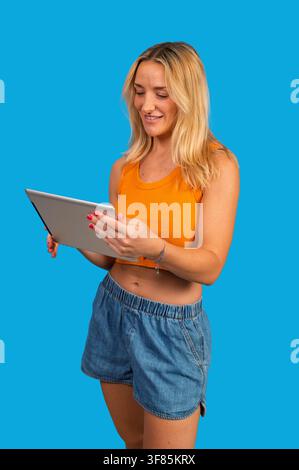 Blonde Frau steht lächelnd und interagiert mit einem digitalen Tablet auf blauem Hintergrund Stockfoto