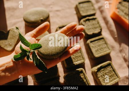 Hand, die eine runde feste Shampoo-Seife mit Minzblatt auf der Handfläche hält, umgeben von handgemachten Seifenstangen in warmem Sonnenlicht Stockfoto