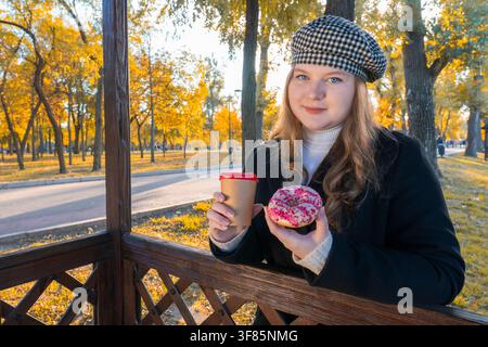 Übergrößenfrau trägt Barett und hält einen Pappkaffeebecher und einen glasierten Donut, während sie an einem Holzpavillon in einem Herbstpark an einem sonnigen Tag lehnt Stockfoto