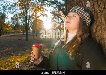 Übergrößen-Frau trägt Barett, lehnt sich an einen Baumstamm, hält einen Pappkaffeebecher und schaut nach oben in einem Herbstpark mit Sonnenlicht-Effekt, Herbstkonzept Stockfoto