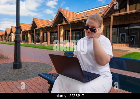 Junge lässige Geschäftsfrau mit Laptop sitzt auf einer Straßenbank und schaut spielerisch über die Sonnenbrille gegen urbane Gebäude Stockfoto