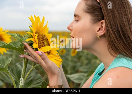 Junge Frau mit geschlossenen Augen lehnt sich zu einer hellen Sonnenblume in einem blühenden Feld, aufgenommen im Seitenprofil als Nahaufnahme mit weichem natürlichem Licht Stockfoto