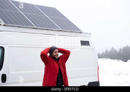 Mittleres Porträt eines glücklichen jungen Mannes, der eine rote Jacke trägt und die Hände auf dem Kopf hat, entspannt vor seinem weißen Wohnmobil mit Solarpanelen in einer s Stockfoto