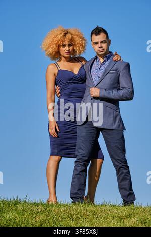 Selbstbewusstes Paar steht auf grünem Gras vor einem klaren blauen Himmel und trägt modische Kleidung Stockfoto