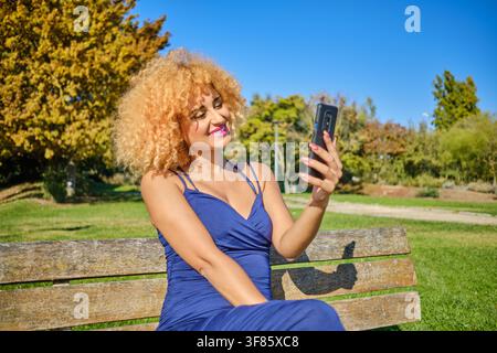 Junge Frau mit lockigem blonden Haar lächelt, während sie im Freien ein Video aufnimmt oder ein Selfie macht Stockfoto