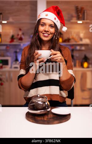 Lächelnde Frau trägt einen Weihnachtsmannhut und posiert in einer mit Weihnachtsschmuck, hellen Lichtern und einem Weihnachtsbaum geschmückten Küche. Stockfoto