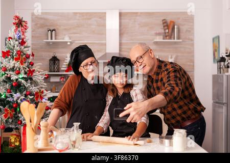 Weihnachts-Selfie fängt zwei Generationen in festlichen Schürzen und Kopfbedeckungen ein, mit einem Großelternteil und einem Kind, die zusammen in der geschmückten Küche lächeln. Stockfoto