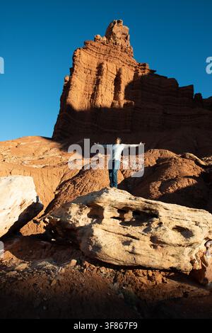 Walker steht auf einem Felsen mit weit ausgebreiteten Armen unter einem riesigen roten Sandsteinturm und feiert die offene Wüstenaussicht. Stockfoto