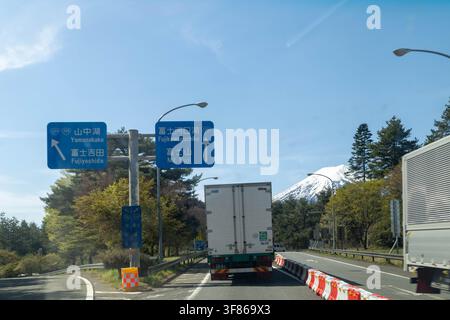 Fujikawaguchiko, Japan - 17. April 2026: Der schneebedeckte Gipfel des Vulkans Fuji und Lastwagen an einer Kreuzung mit Schildern zur Stadt und zum Berg Stockfoto