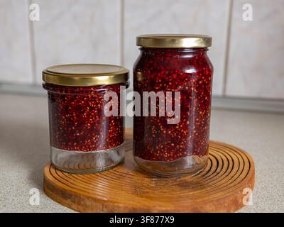 Zwei Gläser mit hausgemachter Walderdbeer-Marmelade stehen auf einem runden Holzschneidebrett vor einem gemütlichen hellen rustikalen Küchenhintergrund Stockfoto