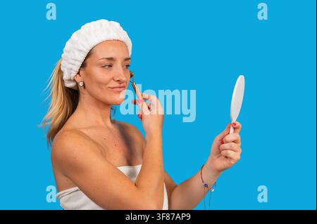 Frau in einem Handtuch und Kopfband, die eine Hautpflegeroutine mit einem Jade-Roller und Spiegel durchführt Stockfoto