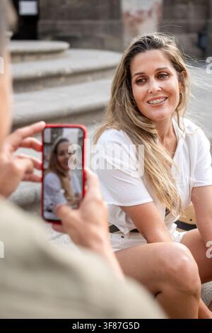 Lächelnde Frau posiert, während ihr Partner mit einem Smartphone ein Foto von ihr macht während eines romantischen Stadturlaubs. Stockfoto