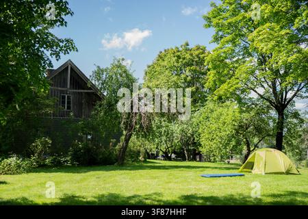 Gelbes Campingzelt und blaue Schlafmatte, aufgestellt auf einem grasbewachsenen Gehöft Rasen in der Nähe von belaubten Obstbäumen, repräsentierend Freizeit im Freien und Ökotourismus Stockfoto