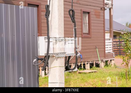 Russland, Sankt Petersburg, 23.08.2025 Stromleitungen und Zähler auf Betonpfeiler mit braunem Holzhaus im Hintergrund. Energieinfrastruktur für r Stockfoto