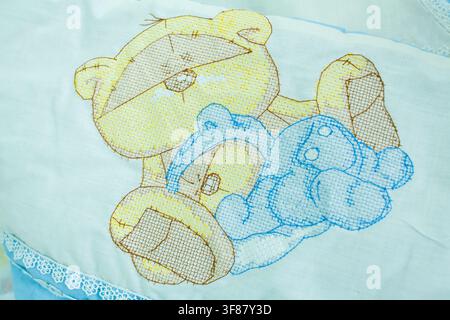 Eine Nahaufnahme zeigt zarte bunte Kreuzstichstickerei niedlicher Teddybären auf einer weichen weißen Babydecke mit Spitzenbesatz. Stockfoto