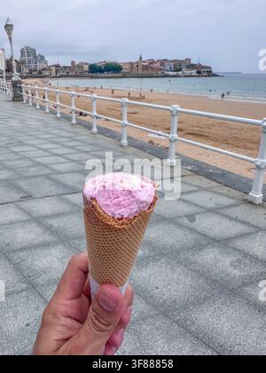 Eine Hand hält eine Eiswaffel mit einer rosa Kugel auf der Strandpromenade von San Lorenzo in Gijón, Asturien, Spanien Stockfoto