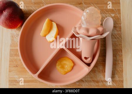 Oberansicht eines rosa Silikon-Saugnapf-Tabletts für Babys mit unterteilten Obstpürees, Apfelscheiben, Nibbler und Löffel auf einem Holzhintergrund. Ideal für Baby Stockfoto
