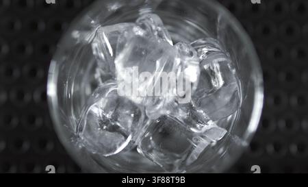 Eiskubeln füllen ein Glas mit erfrischender Kälte, bereiten ein kühles Getränk auf einer Barunterlage vor, Makrodetail von oben Stockfoto