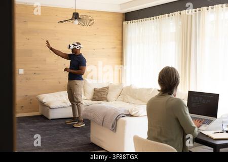 Paar, Mann mit VR-Headset, der im Wohnzimmer greift, während Frau am Laptop programmiert, Kopierraum. Virtual, Interaktion, Fernarbeit, Zuhause, minimalistisch Stockfoto