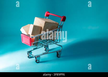 Ein Mini-Einkaufswagen gefüllt mit schlichten Holzblöcken auf einer zyanblauen Oberfläche, die Warenlieferung und E-Commerce-Transaktionen darstellen. Hochwertiges Foto Stockfoto