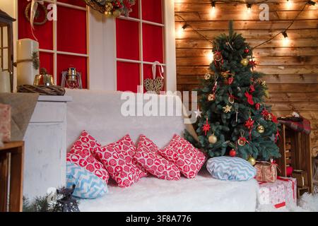 Frohe Weihnachten, Neujahr Wohnzimmer-Innenraum mit Kissen auf dem Sofa, verpackten Geschenkkartons auf dem Teppich, geschmücktem Wintertannenbaum, Kerzen, Kranz auf Rot Stockfoto